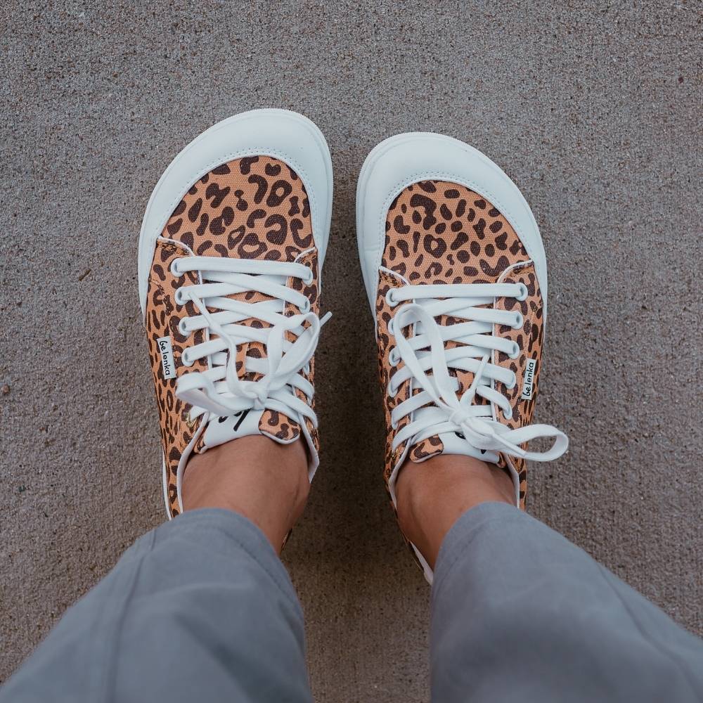 Leopard print Be Lenka Rebound sneakers with white laces, microfiber toe guards, heel accents, and rubber soles. Both shoes are shown from above on a dark skinned woman wearing wide-leg, cropped, grey linen pants standing on cement. #color_leopard