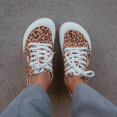 Leopard print Be Lenka Rebound sneakers with white laces, microfiber toe guards, heel accents, and rubber soles. Both shoes are shown from above on a dark skinned woman wearing wide-leg, cropped, grey linen pants standing on cement. #color_leopard