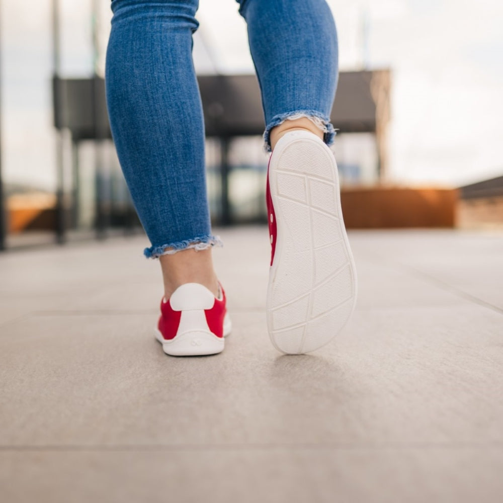 Red Be Lenka Rebound sneakers with white laces, microfiber toe guards, heel accents, and rubber soles. Both shoes are shown from behind on a woman wearing skinny blue jeans with her right toes resting on pavement at the left foot heel showing the right sole with an urban setting in the background. #color_red-white