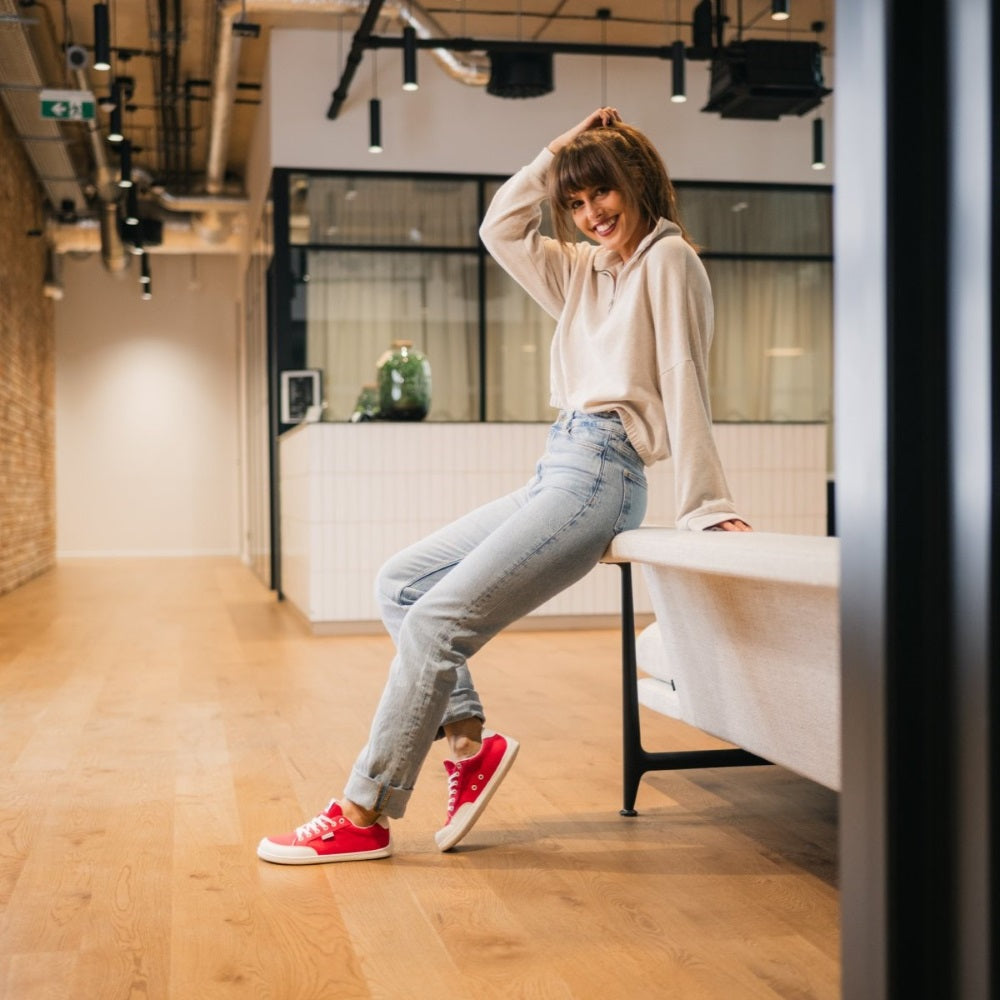 Red Be Lenka Rebound sneakers with white laces, microfiber toe guards, heel accents, and rubber soles. Both shoes are facing left on a woman wearing light-wash, rolled blue jeans and an off white 3/4 zip sweater leaning on a couch with her right toes resting on floor behind the left foot with a modern office setting in the background. #color_red-white