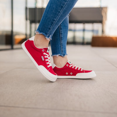 Red Be Lenka Rebound sneakers with white laces, microfiber toe guards, heel accents, and rubber soles. Both shoes are facing right on a woman wearing skinny blue jeans with her right toes resting on pavement at the left foot heel with an urban setting in the background. #color_red-white