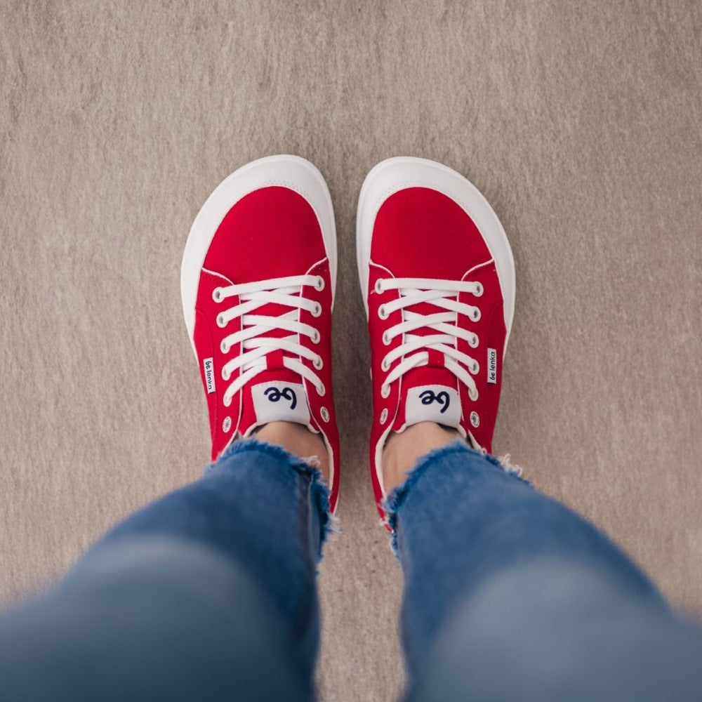 Red Be Lenka Rebound sneakers with white laces, microfiber toe guards, heel accents, and rubber soles. Both shoes are shown from above on a woman wearing skinny blue jeans standing on a grey floor. #color_red-white