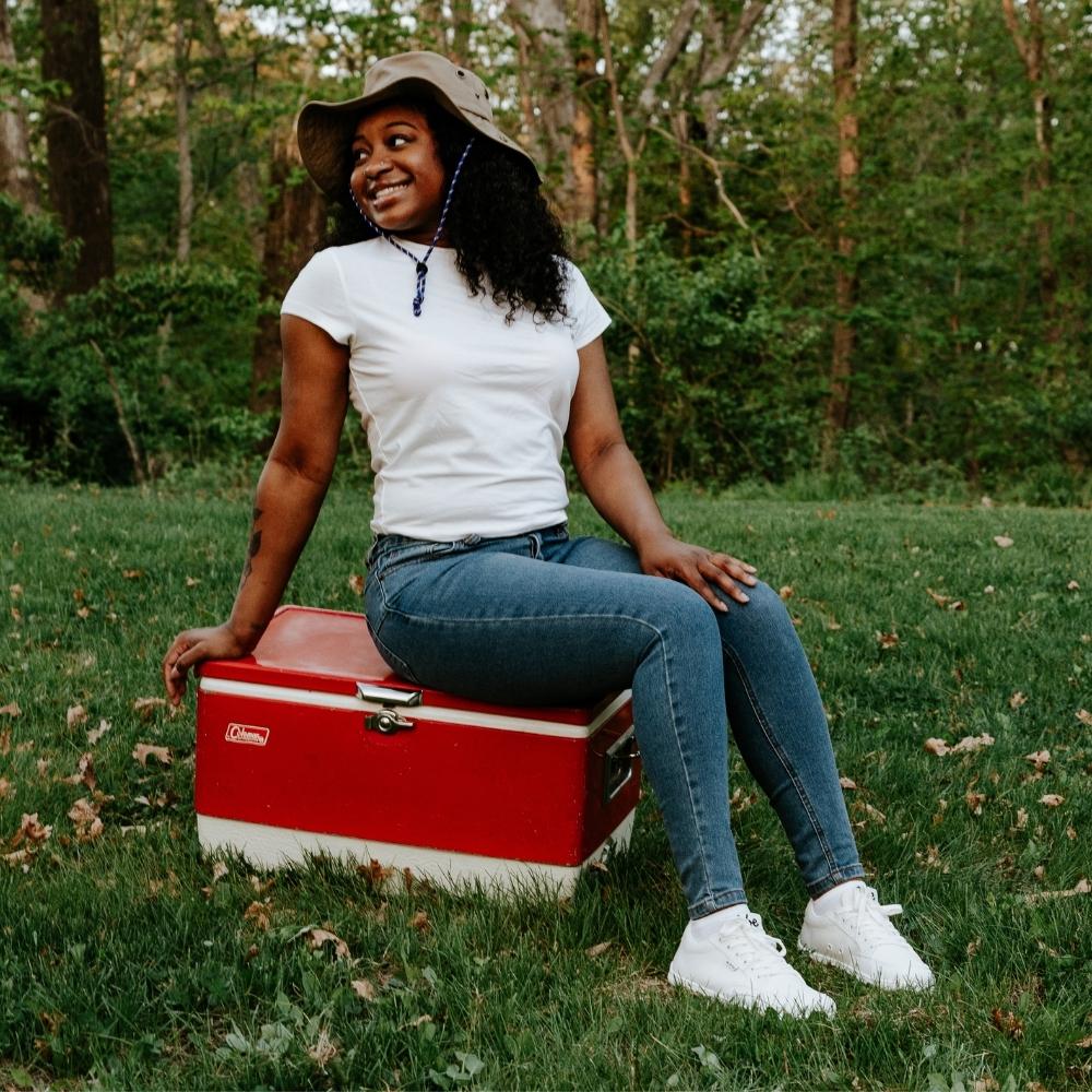 White Be Lenka Rebound sneakers with white laces, microfiber toe guards, heel accents, and rubber soles. Both shoes are shown facing diagonally right on a dark skinned woman with long, curly, black hair sitting on a red cooler in a grassy field  in the woods wearing medium wash skinny jeans, a white shirt, and a fishing hat. #color_all-white