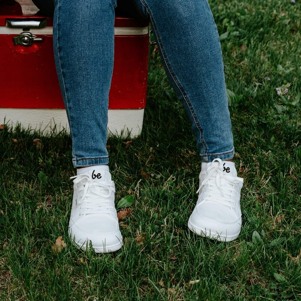 White Be Lenka Rebound sneakers with white laces, microfiber toe guards, heel accents, and rubber soles. Both shoes are shown from the front on a dark skinned woman wearing medium wash skinny jeans sitting on a red cooler in the grass. #color_all-white