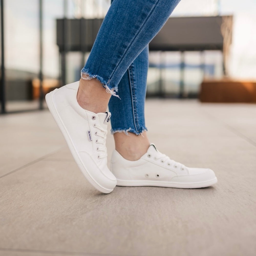 All white Be Lenka Rebound sneakers with white laces, microfiber toe guards, heel accents, and rubber soles. Both shoes are shown facing right on a person wearing torn-edge, blue skinny jeans with the right toe propped on the ground behind. Person is standing on tile floor with an urban background. #color_all-white
