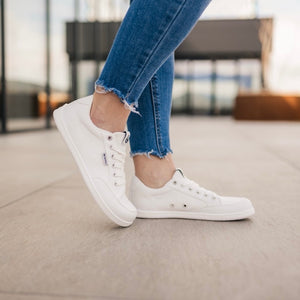 All white Be Lenka Rebound sneakers with white laces, microfiber toe guards, heel accents, and rubber soles. Both shoes are shown facing right on a person wearing torn-edge, blue skinny jeans with the right toe propped on the ground behind. Person is standing on tile floor with an urban background. #color_all-white