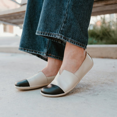 Light beige leather Be Lenka Rosaluna leather loafers with a v cut at the top of the foot, black leather toe caps, and light beige rubber soles. Both shoes are shown facing diagonally left on a light-skinned woman wearing long, medium-wash, loose jeans sitting on a sidewalk bench. #color_light-beige