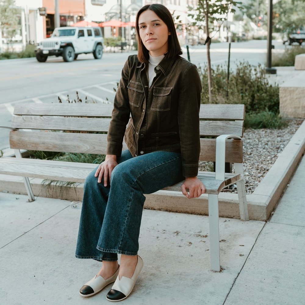 Light beige leather Be Lenka Rosaluna leather loafers with a v cut at the top of the foot, black leather toe caps, and light beige rubber soles. Both shoes are shown facing diagonally left on a light-skinned woman with short brown hair wearing a beige turtle-neck, a brown jean jacket buttoned up, and long, medium-wash, loose jeans sitting on a sidewalk bench in an urban setting. #color_light-beige