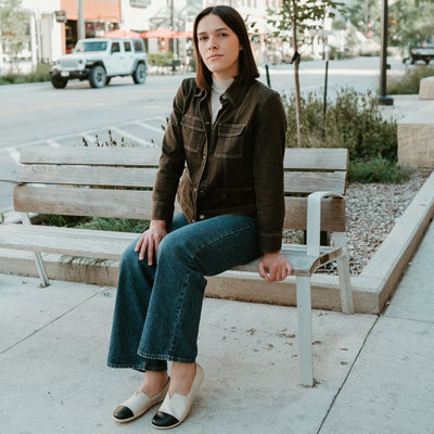 Light beige leather Be Lenka Rosaluna leather loafers with a v cut at the top of the foot, black leather toe caps, and light beige rubber soles. Both shoes are shown facing diagonally left on a light-skinned woman with short brown hair wearing a beige turtle-neck, a brown jean jacket buttoned up, and long, medium-wash, loose jeans sitting on a sidewalk bench in an urban setting. #color_light-beige