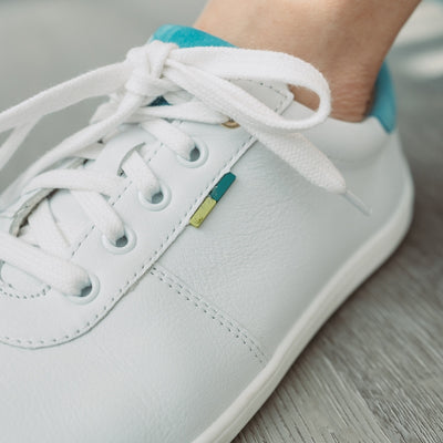 A photo of Be Lenka Royale sneakers made with a leather upper and a rubber sole. The sneakers are a white color and the area around the heel is light blue, they have small yellow and blue tag on the side. One sneaker is shown up close on foot to show the detail of laces and color against a grey wood floor. #color_white-blue