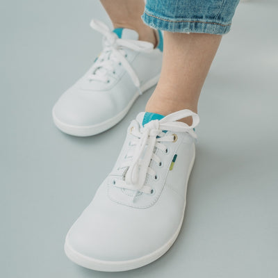 A photo of Be Lenka Royale sneakers made with a leather upper and a rubber sole. The sneakers are a white color and the area around the heel is light blue, they have small yellow and blue tag on the side. A woman is shown from mid-leg down standing on a white floor, she is wearing the mid-wash jeans and the Royale sneakers. #color_white-blue