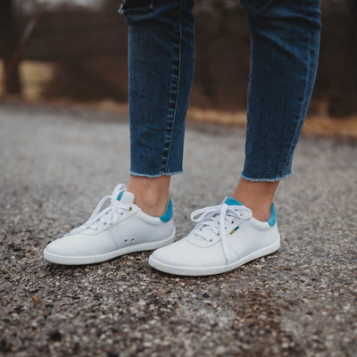 A photo of Be Lenka Royale sneakers made with a leather upper and a rubber sole. The sneakers are a white color and the area around the heel is light blue, they have small yellow and blue tag on the side. A woman is shown facing to the left standing on pavement from knee down wearing dark-wash jeans and the Royale sneakers. #color_white-blue