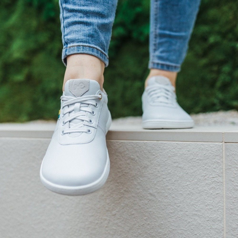A photo of Be Lenka Royale sneakers made with a leather upper and a rubber sole. The sneakers are a white color and the area around the heel is beige, they have small yellow and blue tag on the side. Both shoes are shown from the front on a woman's feet with a view of her shins down. The woman is wearing blue skinny  jeans and is standing on a white rock wall with greenery in the background. #color_white-beige