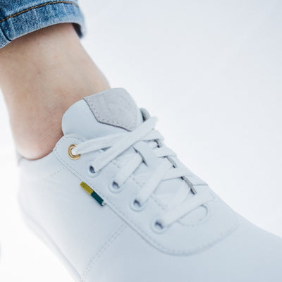 A photo of Be Lenka Royale sneakers made with a leather upper and a rubber sole. The sneakers are a white color and the area around the heel is beige, they have small yellow and blue tag on the side. The right shoe is shown on a woman's foot with a close up view of the top of the shoe on a white background. #color_white-beige