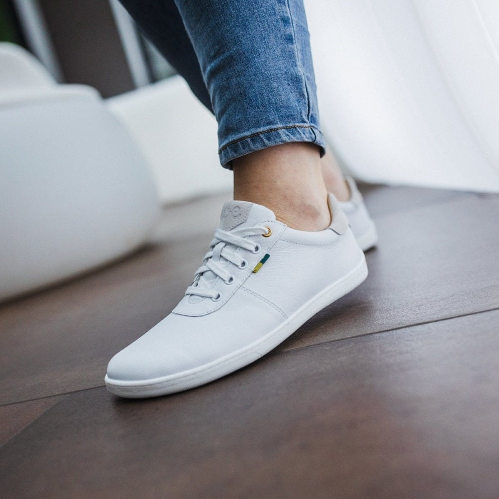 A photo of Be Lenka Royale sneakers made with a leather upper and a rubber sole. The sneakers are a white color and the area around the heel is beige, they have small yellow and blue tag on the side. Both shoes are shown from the left side on a woman's feet with a view of her shins down. The woman is wearing blue skinny  jeans and is standing on a brown floor. #color_white-beige