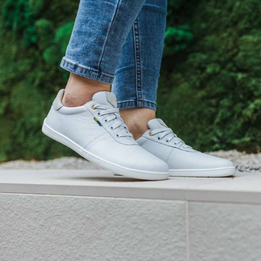 A photo of Be Lenka Royale sneakers made with a leather upper and a rubber sole. The sneakers are a white color and the area around the heel is beige, they have small yellow and blue tag on the side. Both shoes are shown from the right side on a woman's feet with a view of her shins down. The woman is wearing blue skinny  jeans and is standing on a white rock wall with greenery in the background. #color_white-beige
