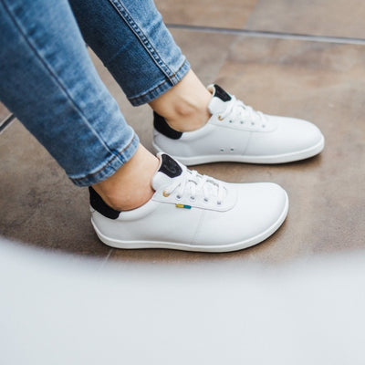 A photo of Be Lenka Royale sneakers made with a leather upper and a rubber sole. The sneakers are a white color and the area around the heel is black, they have small yellow and blue tag on the side. Both sneakers are shown from the right on a woman's feet with a view of her knees down. The woman is wearing blue skinny jeans and is standing on brown stone. #color_white-black