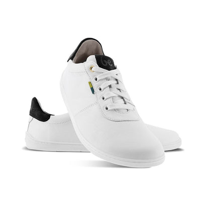 A photo of Be Lenka Royale sneakers made with a leather upper and a rubber sole. The sneakers are a white color and the area around the heel is black, they have small yellow and blue tag on the side. Both shoes are shown, the left shoe is behind the right. The right shoes heel is leaning up against the left shoe showing the right shoe from the front facing down against a white background. #color_white-black