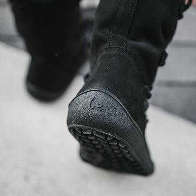A photo of Be Lenka Adult Snowfox boots in all black. Black leather goes around the back with black satin over the top and front, and black soles. Laces are widely connected from side to side starting at the ball of the foot and going until the top of the boot. Both boots are shown from the back on a woman's feet with a close up of the right heel. The woman is walking on a paved sidewalk. #color_black