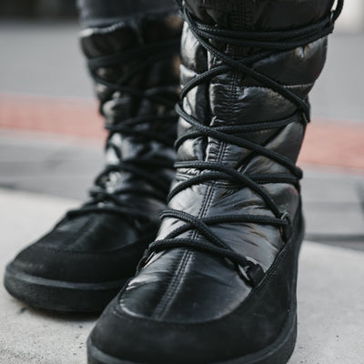 A photo of Be Lenka Adult Snowfox boots in all black. Black leather goes around the back with black satin over the top and front, and black soles. Laces are widely connected from side to side starting at the ball of the foot and going until the top of the boot. Both boots are shown from the front on a woman's feet to show the laces.The woman is wearing dark gray skinny jeans tucked into the boots.  #color_black