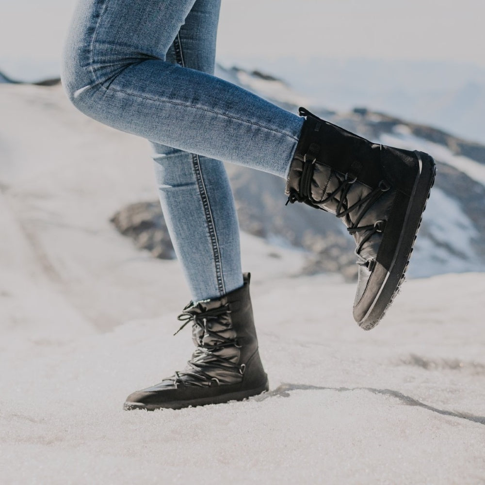 A photo of Be Lenka Adult Snowfox boots in all black. Black leather goes around the back with black satin over the top and front, and black soles. Laces are widely connected from side to side starting at the ball of the foot and going until the top of the boot. Both boots are shown facing left on a woman wearing light skinny jeans tucked into the boots standing on a snow covered mountain. #color_black