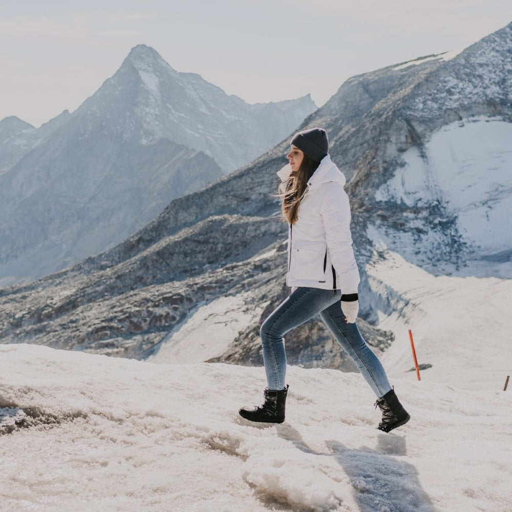 A photo of Be Lenka Adult Snowfox boots in all black. Black leather goes around the back with black satin over the top and front, and black soles. Laces are widely connected from side to side starting at the ball of the foot and going until the top of the boot. Both boots are shown facing left on a woman climbing a snow covered mountain wearing light skinny jeans tucked into the boots, a puffy white coat, white hand warmers, and a black hat. #color_black