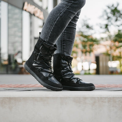 A photo of Be Lenka Adult Snowfox boots in all black. Black leather goes around the back with black satin over the top and front, and black soles. Laces are widely connected from side to side starting at the ball of the foot and going until the top of the boot. Both boots are shown from the right side on a woman's feet who is walking on a paved sidewalk with a building and a tree with green leaves in the background. The woman is wearing dark gray skinny jeans tucked into the boots. #color_black