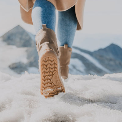 A photo of Be Lenka Adult Snowfox boots in all black. Brown leather goes around the back with champagne colored satin over the top and front, and black soles. Laces are widely connected from side to side starting at the ball of the foot and going until the top of the boot. Both shoes are shown from the back. Left boot sole is shown in the foreground on a woman wearing light wash skinny jeans and a long tan coat standing on a snow covered mountain. Right boot is in the background. #color_champagne-brown