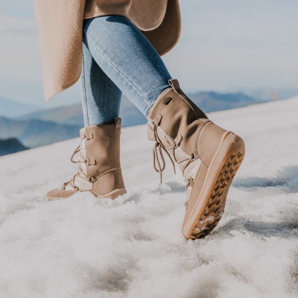 A photo of Be Lenka Adult Snowfox boots in all black. Brown leather goes around the back with champagne colored satin over the top and front, and black soles. Laces are widely connected from side to side starting at the ball of the foot and going until the top of the boot. Both shoes are shown from the back left diagonal with the left boot in the foreground showing the side and sole on a woman wearing light wash skinny jeans and a long tan coat standing on a snow covered mountain. #color_champagne-brown