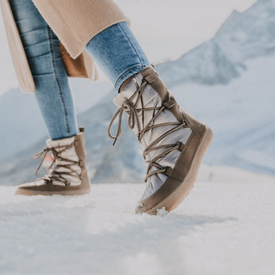 A photo of Be Lenka Adult Snowfox boots in all black. Brown leather goes around the back with champagne colored satin over the top and front, and black soles. Laces are widely connected from side to side starting at the ball of the foot and going until the top of the boot. Both shoes are shown facing left with the left boot in the foreground on a woman wearing light wash skinny jeans and a long tan coat standing on a snow covered mountain. #color_champagne-brown