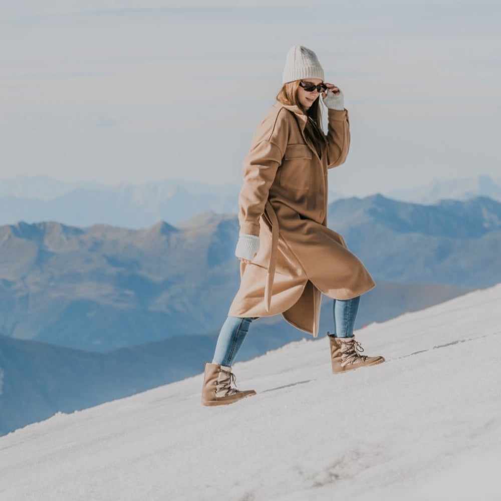 A photo of Be Lenka Adult Snowfox boots in all black. Brown leather goes around the back with champagne colored satin over the top and front, and black soles. Laces are widely connected from side to side starting at the ball of the foot and going until the top of the boot. Both shoes are shown facing right on a woman wearing light wash skinny jeans tucked into the boots, a long tan coat, and white hand warmers and hat climbing up a snow covered mountain. #color_champagne-brown