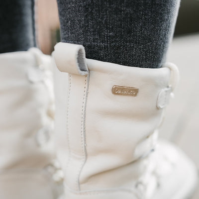 A photo of Be Lenka Adult Snowfox boots in Pearl White. Pearl white leather goes around the back with pearly white satin over the top and front, and white soles. Laces are widely connected from side to side starting at the ball of the foot and going until the top of the boot. The top of the right boot is shown from the back right on a woman's foot to show the leather upper. The woman is wearing dark gray skinny jeans tucked into the boots. #color_pearl-white