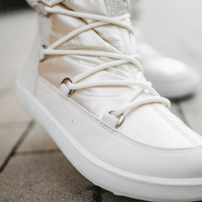 A photo of Be Lenka Adult Snowfox boots in Pearl White. Pearl white leather goes around the back with pearly white satin over the top and front, and white soles. Laces are widely connected from side to side starting at the ball of the foot and going until the top of the boot. The laces on the right boot are shown from the front right on a woman's foot on a paved sidewalk. #color_pearl-white