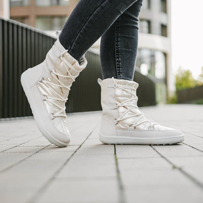 A photo of Be Lenka Adult Snowfox boots in Pearl White. Pearl white leather goes around the back with pearly white satin over the top and front, and white soles. Laces are widely connected from side to side starting at the ball of the foot and going until the top of the boot. Both boots are shown from the right on a woman's feet while she is walking on a paved sidewalk with a black fence and a building in the background. The woman is wearing dark gray skinny jeans tucked into the boots. #color_pearl-white