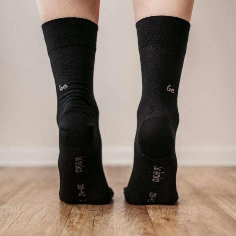 A photo of Be Lenka crew cut socks in black. Boths socks are show here on feet from the back with the left foot bending forward showing the bottom of the sock atop a brown wooden floor. #color_black