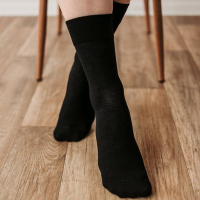 A photo of Be Lenka crew cut socks in black. Boths socks are show here on feet from the front with the right foot crossed over the left atop a brown wooden floor with chair legs in the background. #color_black