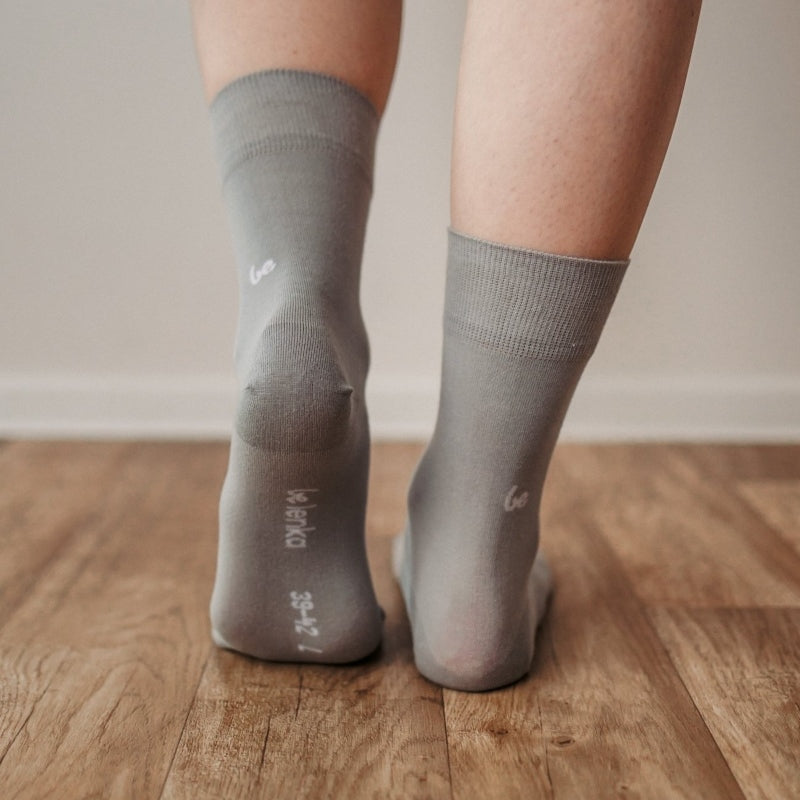 A photo of Be Lenka crew cut socks in grey. Boths socks are show here on feet from the back with the left foot bending forward showing the bottom of the sock atop a brown wooden floor. #color_grey