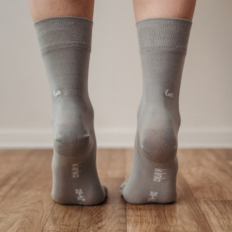 A photo of Be Lenka crew cut socks in grey. Boths socks are show here on feet from the back with the model standing on their toes showing the bottom of the sock atop a brown wooden floor. #color_grey