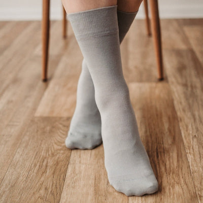 A photo of Be Lenka crew cut socks in grey. Boths socks are show here on feet from the front with the right foot crossed over the left atop a brown wooden floor with chair legs in the background. #color_grey
