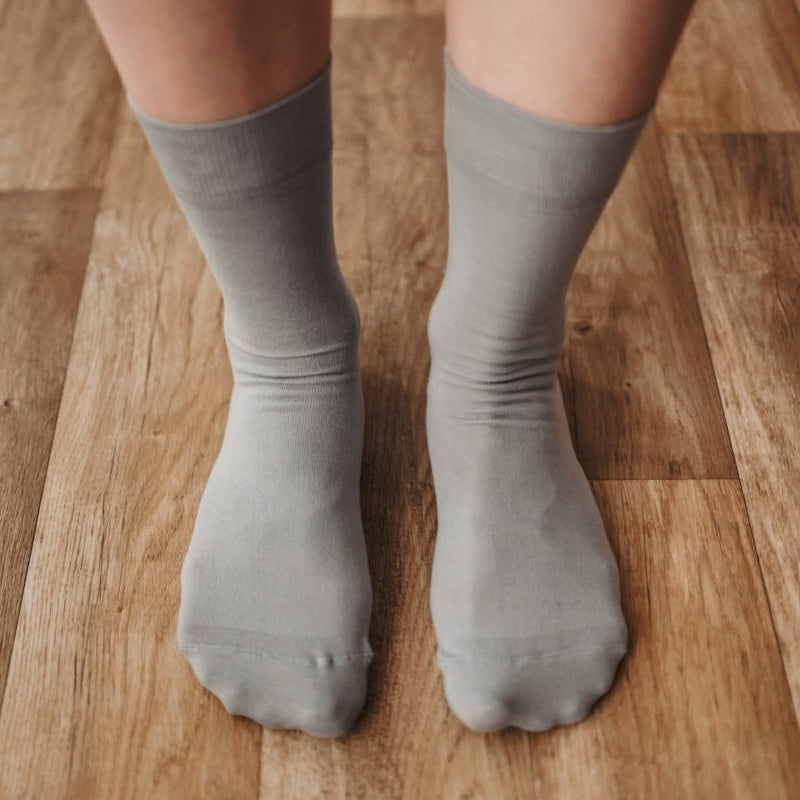 A photo of Be Lenka crew cut socks in grey. Boths socks are show here on feet from the top down atop a brown wooden floor. #color_grey