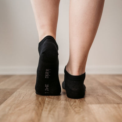 A photo of Be Lenka low cut socks in black. Boths socks are show here on feet from the back with the left foot bending forward showing the bottom of the sock atop a brown wooden floor. #color_black