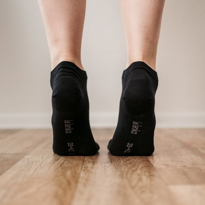 A photo of Be Lenka low cut socks in black. Boths socks are show here on feet from the back with the model standing on their toes showing the bottom of the sock atop a brown wooden floor. #color_black