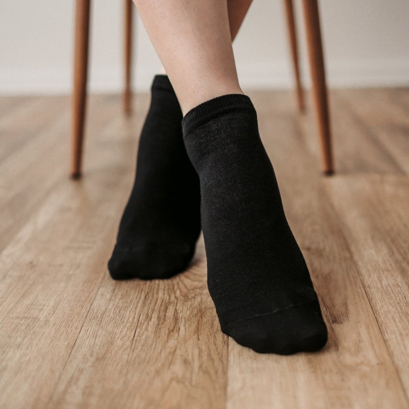 A photo of Be Lenka low cut socks in black. Boths socks are show here on feet from the front with the right foot crossed over the left atop a brown wooden floor with chair legs in the background. #color_black