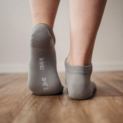A photo of Be Lenka low cut socks in grey. Boths socks are show here on feet from the back with the left foot bending forward showing the bottom of the sock atop a brown wooden floor. #color_grey
