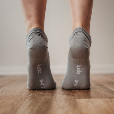 A photo of Be Lenka low cut socks in grey. Boths socks are show here on feet from the back with the model standing on their toes showing the bottom of the sock atop a brown wooden floor. #color_grey