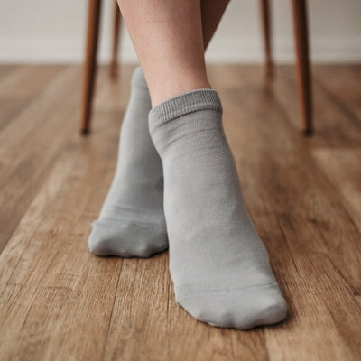 A photo of Be Lenka low cut socks in grey. Boths socks are show here on feet from the front with the right foot crossed over the left atop a brown wooden floor with chair legs in the background. #color_grey