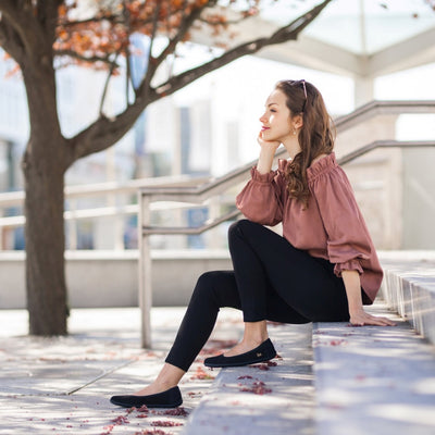 A photo of the Be Lenka Sophie flats made from a leather upper and a microfiber interior. The flats are black in color with black rubber soles and are a simple ballerina flat design. Both shoes are shown facing left on a light-skinned woman with brown hair. She is wearing black, cropped dress pants and a loose, silky, off-the-shoulder mauve shirt sitting on stairs with an urban setting and a tree in the background. #color_matte-black