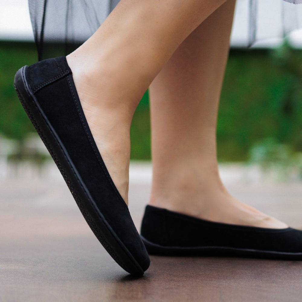 A photo of the Be Lenka Sophie flats made from a leather upper and a microfiber interior. The flats are black in color with black rubber soles and are a simple ballerina flat design. Both shoes are shown from the right side on a woman's feet with a view of her shins down. The woman is walking on a brown floor with her right foot behind her left.  #color_matte-black