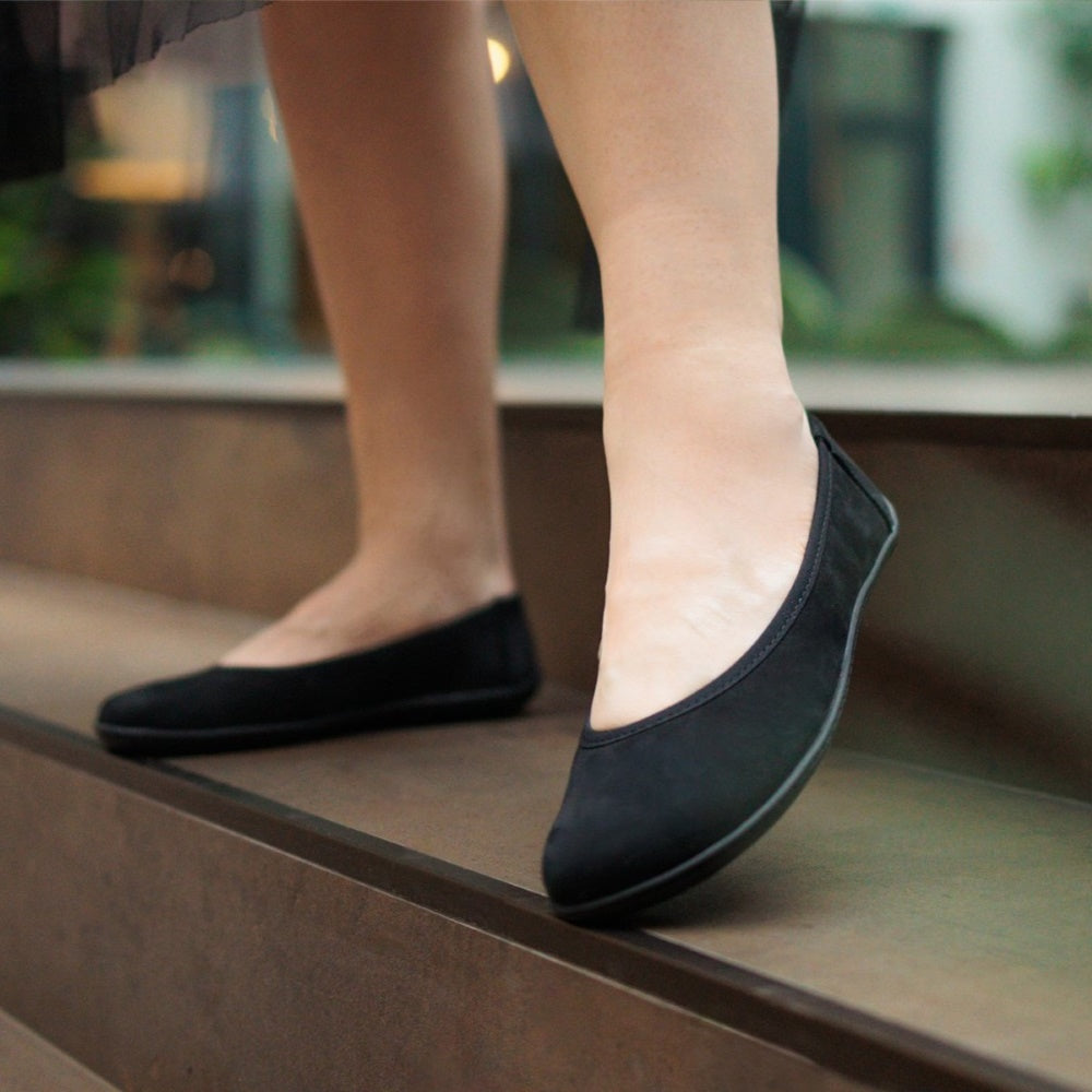 A photo of the Be Lenka Sophie flats made from a leather upper and a microfiber interior. The flats are black in color with black rubber soles and are a simple ballerina flat design. Both shoes are shown from the front on a woman's feet with a view of her shins down. The woman is standing on a brown step. #color_matte-black