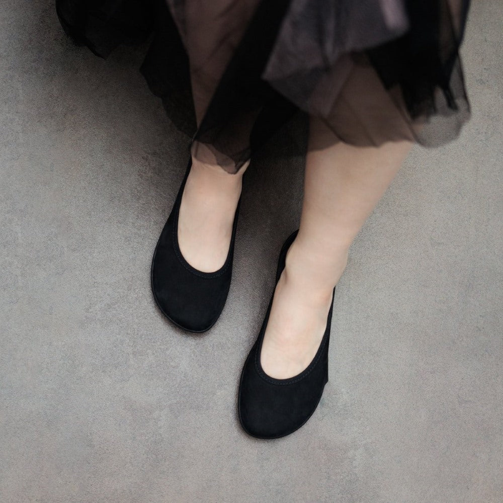 A photo of the Be Lenka Sophie flats made from a leather upper and a microfiber interior. The flats are black in color with black rubber soles and are a simple ballerina flat design. Both shoes are shown from above on a woman's feet with a view of her knees down. The woman is wearing a black tulle skirt and is standing on a gray cement floor. #color_matte-black